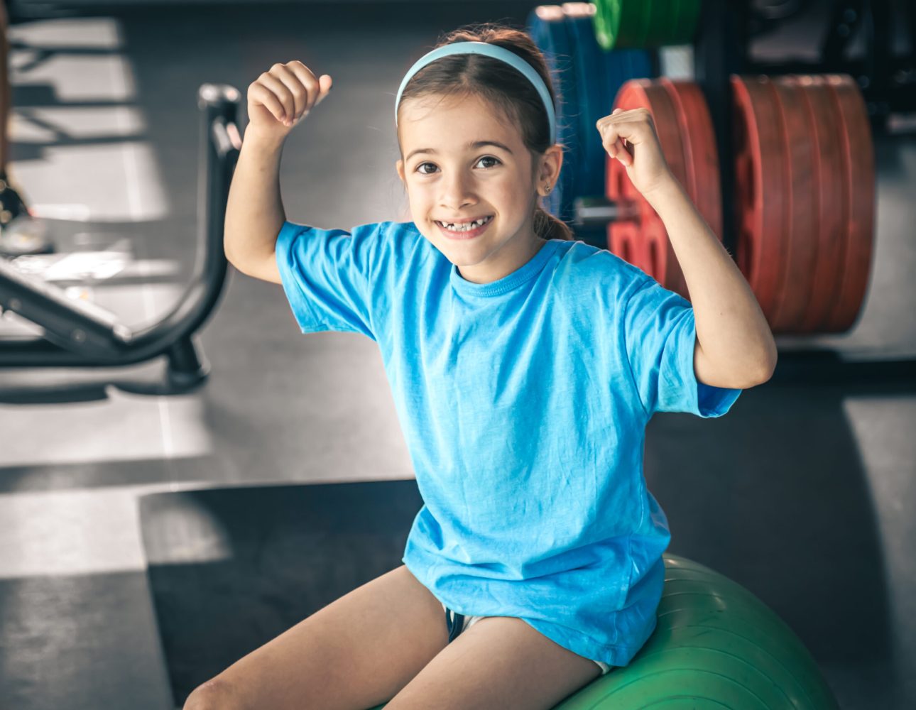 Charming little girl is shows her biceps, working out at gym. Charming little girl is shows her biceps and smiling while working out at gym.